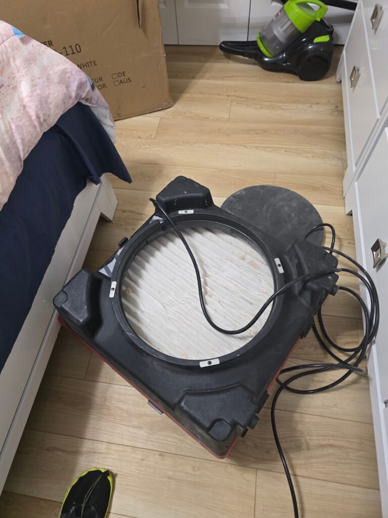 A portable air scrubber with an exposed filter rests on a light wood floor next to a bed.