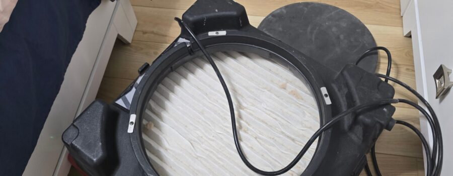 A portable air scrubber with an exposed filter rests on a light wood floor next to a bed.