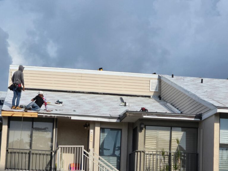 Two roofers work on installing underlayment on the roof of a residential building under an overcast sky.