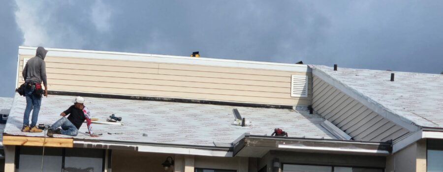 Two roofers work on installing underlayment on the roof of a residential building under an overcast sky.