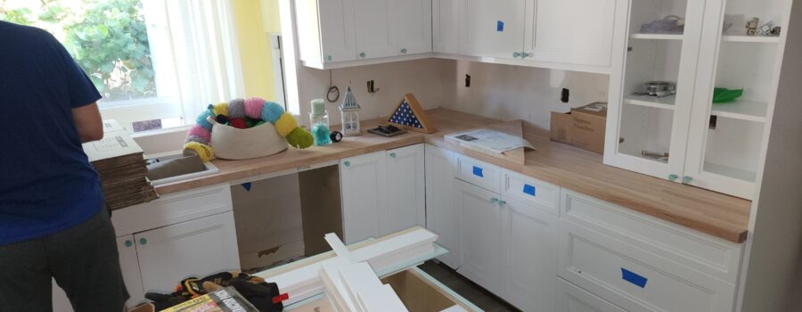 A kitchen undergoes renovation with newly installed white cabinets, light wood countertops, and construction supplies scattered across the workspace.